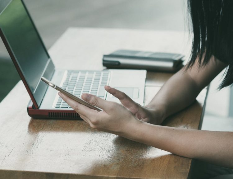A young woman multitasking with her smartphone and laptop at a table indoors.