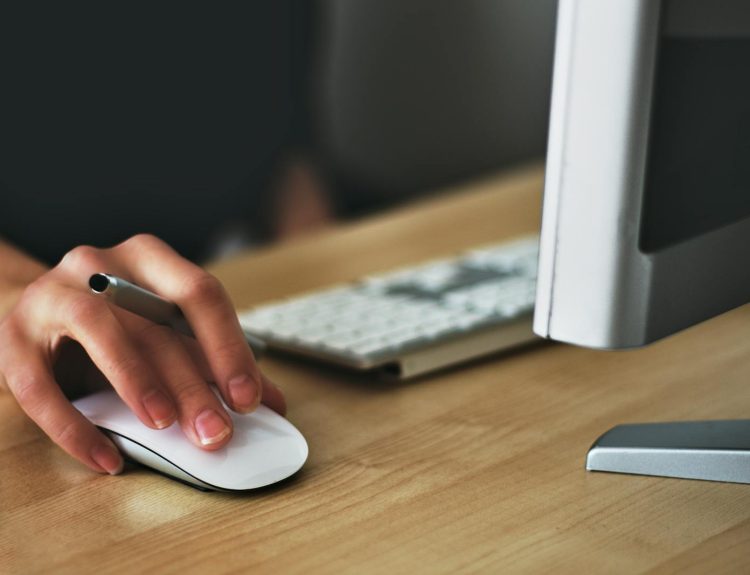 A hand using a wireless mouse at a modern desk setup with a computer and keyboard.