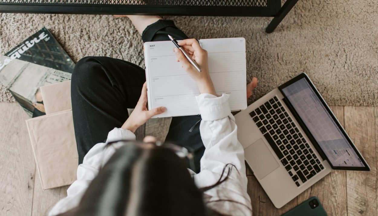 Top view of anonymous woman in casual wear sitting on floor with laptop and smartphone and creating plan on notebook while resting during break in modern living room