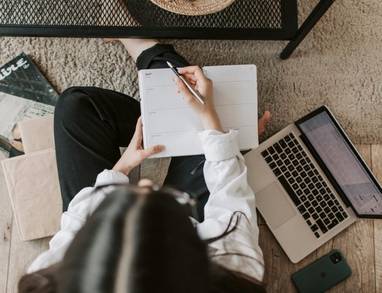Top view of anonymous woman in casual wear sitting on floor with laptop and smartphone and creating plan on notebook while resting during break in modern living room