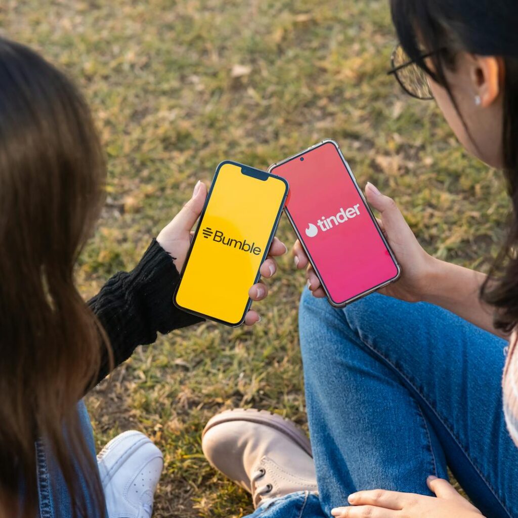 Two women using Bumble and Tinder apps on smartphones in a park setting.