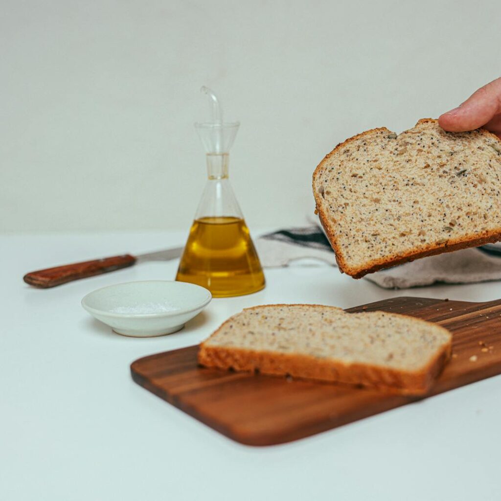 A hand holding a slice of wheat bread with olive oil, knife, and saucer in view, ideal for stock photography.